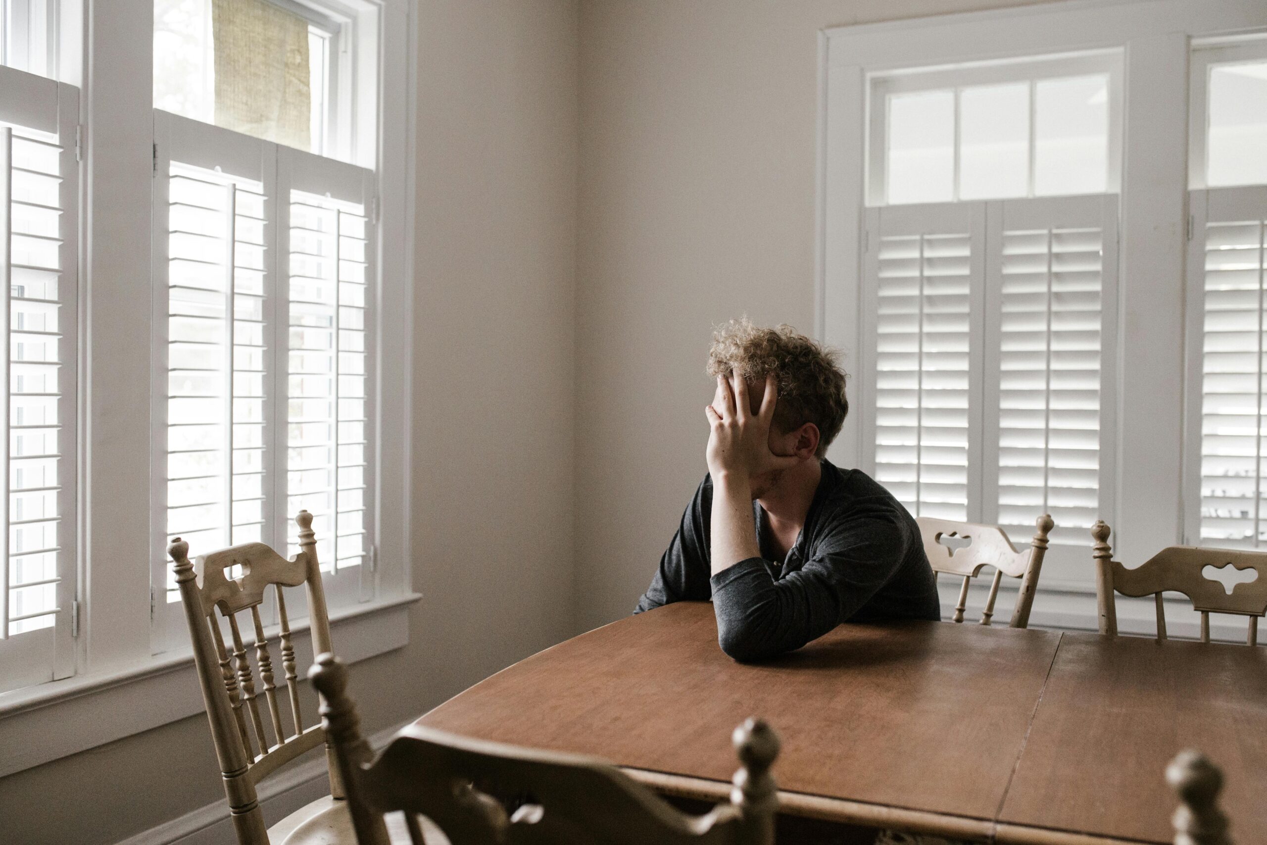 Homem sentado em uma mesa com cadeiras de madeira, o espaço é branco e bem iluminado, porém vazio. Este homem está com a mão no rosto, em posição que sugere que está incomodado e pensativo. Foto por Andrew (pexels).